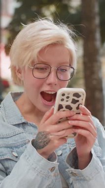 Kafkasyalı genç bir kadın cep telefonuyla kutlama yapıyor iyi haber kazanıyor, piyango büyük ikramiyesi kazanıyor, açık havada dağıtıyor. Şehir caddesinde duran mutlu sarışın bayan turist. Kasaba yaşam tarzları.