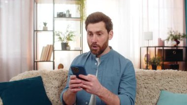 Genç yetişkin bir adam evde oturmuş, akıllı telefonuyla sosyal medyaya odaklanmış bir ifadeyle bakıyor. Kanepedeki beyaz adam beklenmedik online içerik keşfettikten sonra aniden kaşlarını kaldırdı.