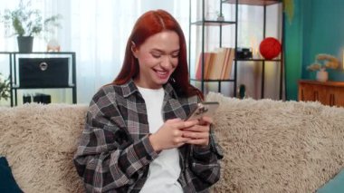 Genç ve mutlu bir kadın evde akıllı telefondan mesaj atıyor. Odaklanıp gülüyor ve kurye uygulamasında sohbet ediyor. Kanepedeki beyaz saçlı kız eğlenceli sohbetler yapıyor. Komik mesajlara sevinçle tepki veriyor.