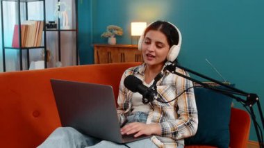 Evde dizüstü bilgisayarda çalışan, kulaklık takan, profesyonel stüdyo mikrofonuna konuşan Hintli genç bir kadın. Çevrimiçi yayına odaklanmış Arap mutlu kız aktif olarak podcast yayınlıyor veya kaydediyor