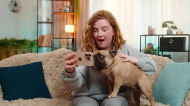 Mutlu Kafkasyalı kızıl kadın akıllı telefondan komik videolar izleyip kanepede oturmuş kahkahalarla gülüyor. Pug dog ekranı merakla izliyor. Huzurlu ortamda paylaşılan neşeli ana tepki veriyor..