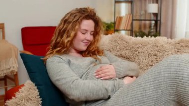 Hamile genç bir kadın, iki eliyle karnına hafifçe dokunarak, rahat ve huzurlu bir anın tadını çıkarıyor. Kız sükuneti kucaklıyor, sevgiyi hissediyor ve gelecekteki bebekle, annelikle bağ kuruyor.