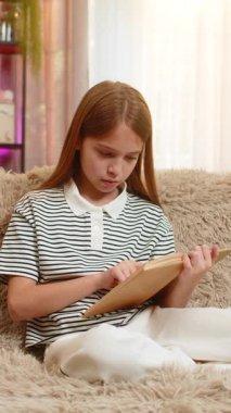 Genç kız kanepede otururken büyüleyici bir kitap okuyor, hikayeyi incelerken ilgiyle ve sevinçle gülümsüyor. Okul çocuğu sessiz okumaları sever, rahat ev ortamında mutlu olur.