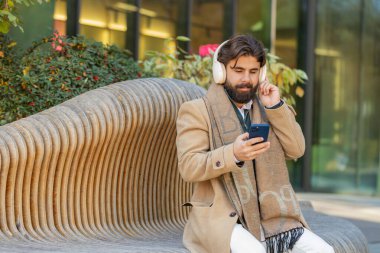 Kablosuz kulaklıkla dans eden mutlu, rahat Hintli genç işadamının portresi, akıllı telefon dansında enerjik disko müziği dinlemek. Lübnanlı sakallı adam şehir merkezindeki bankta oturuyor.