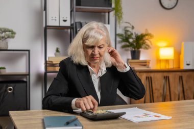 İş yerindeki finans raporları için hesap yapan üzgün bir muhasebeci kadın. Ciddi bir yaşlı kadın büyükanne, hesap makinesi kullanıyor. Ofis masasında oturuyor. Muhasebe kavramı.