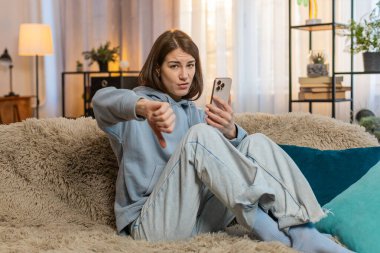 Hoşlanmamak. Kafkasyalı kadın akıllı telefonu evdeki kanepede otururken kullanıyor. İşaret işareti işareti yapıyor, hoşnutsuzluğunu dile getiriyor, tatminsiz kötü işleri tasvip ediyor. Memnun olmayan kız başını sallar..