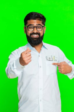 Hintli adam kendine güvenen yüzüyle ve parmaklarıyla para jesti yaparak kişisel zenginliğini finansal başarıyla ifade ediyor. Arap adam yeşil krom anahtar arka planda izole nakit sinyal refah ruh hali gösteriyor