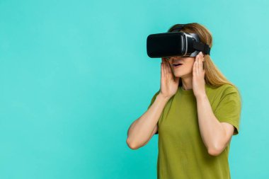 Orta yaşlı kadın VR kulaklık takıyor ve dikkatlice etrafa bakıyor, meraklı odaklı yüzüyle dijital çevreyi keşfediyor. Mavi arka planda izole edilmiş kız sanal dünyaya ilgi göstererek tepki veriyor