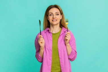 Orta yaşlı bir kadın elinde çatal ve bıçak ile istekli bir yüz ifadesi ile aç ve yemeye hazır olduğunu açıkça gösteriyor. Mavi arkaplanda izole edilmiş bir kız klasik yemek zamanı jestini beklentiyle gösterir