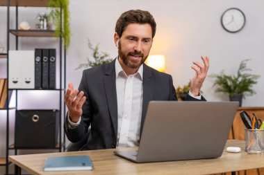 Merkez ofisteki orta yaşlı işadamı kablosuz kulaklık takarak bilgisayar konferansına katılıyor. Masadaki serbest çalışan adam güler ve toplantı sırasında not defterine not alır..