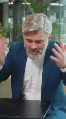 Çatışma Anlaşması 'nda. Olgun kızgın patron iş adamı webcam çevrimiçi video görüşmesi yapıyor, kameraya bakarak iş arkadaşlarıyla konuşuyor. Takım elbiseli beyaz adam modern ofis lobisinde oturuyor..