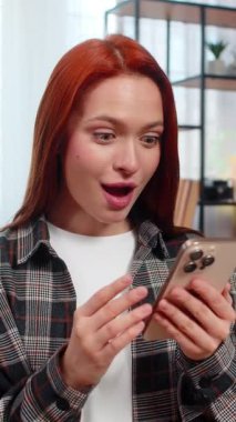 Evde akıllı telefondan güzel haberler okuyan, gülümseyen ve heyecanla el kaldıran genç bir kadın. Kafkasyalı kızıl saçlı kız kanepeye oturmuş neşeli bir başarıyı kutluyor, mutlu, olumlu duygusal bir zafer yaşıyor.