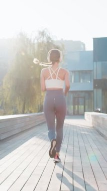 Antrenman maratonunda koşan, kulaklıkla müzik dinleyen aktif sporcu kadının arka görüntüsü. Şehir parkında kardiyo çalışması. Genç kız sağlıklı yaşam tarzını seviyor. Kadın atlet koşucu.