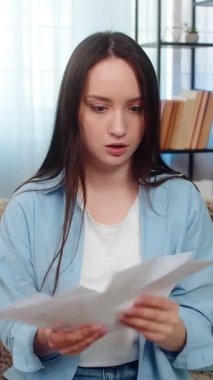 Evdeki sinirli genç kadın kanepeye oturmuş gergin bir şekilde tartışıyor, finansal belgeleri kanepeye fırlatıyor. Kafkasyalı kız borçlar ve ekonomik krizle vurgulandı, mali başarısızlığa duygusal tepki gösterdi