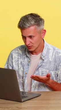 Orta yaşlı bir adam dizüstü bilgisayarla masada oturuyor, megafona bağırıyor sanki duyulmamış gibi saldırgan bir şekilde el kol hareketi yapıyor. Genç adam sarı arka planda online çatışma sırasında öfke ve hayal kırıklığını ifade ediyor