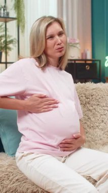 Hamile, orta yaşlı bir kadın karnına sarılıyor kramplardan nefes alıyor karın ağrısı çekiyor ve rahatlık arıyor. Kanepede oturan beyaz bir kız gözleri kapalı, ağrıyı kontrol ediyor ve bebek güvenliği için hızlı bir rahatlama umuyor.