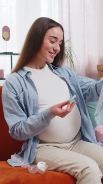 Hamile kadın bebek bezi ve emziğine bakıyor. Müstakbel anne anneliği seviyor, evdeki kanepede oturan şefkatli bebek bezlerine bak. Hamilelik hazırlığı, bebek, doğum konsepti.