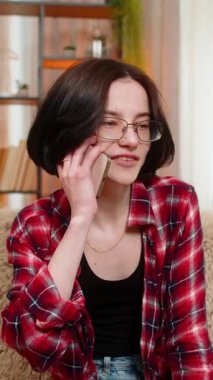 Genç bir kadın, evinde kanepede oturmuş sevinçle gülümseyen ve el hareketlerini etkileyen akıllı telefonuyla konuşuyor. Kız sıcak çevrimiçi sohbetleri sever, arkadaş canlısı tavırları ve olumlu duyguları ifade eder..