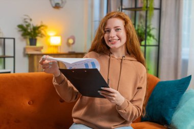 Finansal grafik belgeleriyle çalışan genç kadın muhasebeci, çizelgeler için vergileri hesaplıyor ve kanepedeki ev gelirlerinin grafiklerini çıkarıyor. Kanepede oturan kızıl saçlı, harcama bütçeli yetişkin kız..