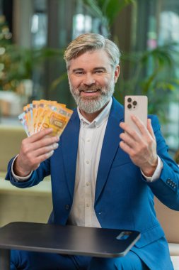 Başarılı, mutlu, olgun bir iş adamı akıllı telefon tutuyor ve ofis lobisinde oturmuş euro banknotlarını sayıyor. İnternette piyango kazanan beyaz sakallı adam. Bütçe hesaplanıyor.