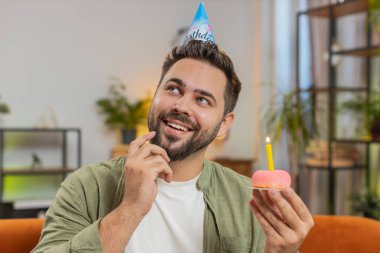 Mutlu adam elinde kek tutan neşeli bir doğum günü şapkası takıyor pastaya mum üflediği için sevinçle tebrik ediyor. Genç esmer adam doğum günü partisini sınıftaki kanepede kutluyor..