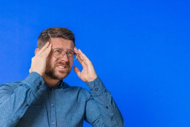 Memnun olmayan orta yaşlı, gözlüklü, baş ağrısı problemini tedavi etmek için şakak ovalayan, migren stresinden muzdarip yaşlı bir adam. Acı içinde kıvranan bir adam, mavi arka planda yüksek tansiyon.