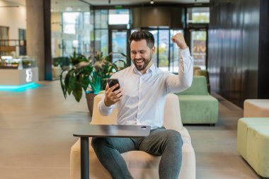 Beyaz gömlekli mutlu genç işadamı piyango ikramiyesi kazanan akıllı telefondan şok geçiriyor. Kariyer başarısı iyi haber. Ofis lobisinde oturan heyecanlı adam..