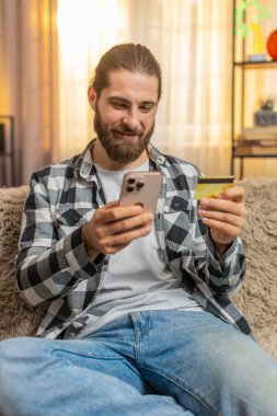 Genç adam evde telefon ve kredi kartıyla oturuyor, çevrimiçi alışverişi sonuçlandırırken toplamı doğruluyor. Kanepede oturan beyaz adam tamam, başarılı ödeme işlemlerine güvenle kafa sallıyor.