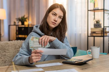 Kanepede oturan genç beyaz kadın faturaları incelerken para sayıyor. Yetişkin kız makbuzları kontrol eder ve masadaki harcamaları ve bütçeyi odaklanmış ifadeyle hesap makinesi kullanır.