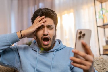 Akıllı telefon kullanan, negatif mesaj okuyan, oturma odasında kanepede oturmaktan rahatsız olan üzgün genç adam şok oldu. Alet sorunu olan Hintli adam üzgün görünüyor, telefonu tutuyor ve kötü habere tepki veriyor..