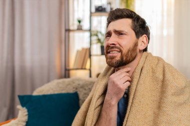 Evdeki genç adam kanepeye oturur, battaniyeye sarınır, öksürür ve acı içinde boğazını tutar. Kafkasyalı adam kaşlarını çatmış, yutkunmaya çalışmış, muhtemelen boğaz ağrısı ya da grip belirtileri göstermiş.