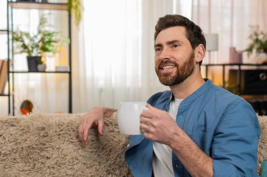 Evdeki adam kanepede oturup dinleniyor, sabahları elinde bir fincan sıcak çay tutarak düşünceli bir şekilde uzaktan bakıyor. Adam huzurlu bir andan hoşlanıyor, rahatlamış hissediyor, muhtemelen derin düşüncelere dalmış.