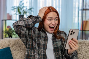 Evde akıllı telefondan güzel haberler okuyan, gülümseyen ve heyecanla el kaldıran genç bir kadın. Kafkasyalı kızıl saçlı kız kanepeye oturmuş neşeli bir başarıyı kutluyor, mutlu, olumlu duygusal bir zafer yaşıyor.