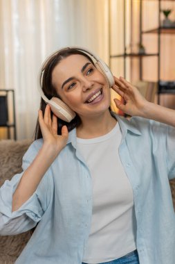 Evde kulaklık takan genç bir kadın ritmik olarak hareket ediyor, müzik dinlerken mutlu bir şekilde dans ediyor. Kanepede oturan beyaz bir kız neşeli duygular, olumlu hisler, eğlenceli bir eğlence..