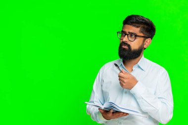 Hintli adam defterine ciddi odaklanmış bir ifadeyle günlük işleri planlar ve yapılacaklar listesi düzenler. Arap adam yeşil krom anahtar arka planda izole edilmiş. Konsantrasyon ve hazırlık gösteriyor.