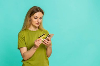 Orta yaşlı bir kadın elinde akıllı telefon ve kredi kartı tutarken sipariş düğmesine tıklayarak dijital ödeme anını özgüvenle gösteriyor. Mavi arka planda yetişkin bir kız. Odaklanmış duygularla internetten mal alıyor..