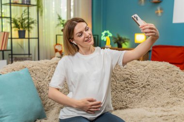 Evde orta yaşlı hamile bir kadın kulaklık takıyor. Akıllı telefon görüşmeleri yapıyor. Merhaba, göbek konuşuyor. Kanepedeki beyaz kız aile sohbetinin keyfini çıkarıyor bebek planları hakkında mutlu bir gülümseme destek veriyor..