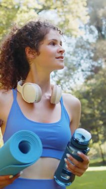 Elinde hasırla genç ve mutlu sporcu kadın parktaki açık hava eğitimine gidiyor. Aktif giyimli, kulaklık takan ve su şişesi kullanan iyimser gülümseyen kız jimnastik eğitimine gidiyor..