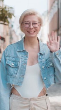 Neşeli genç bayan turist kameraya gülümsüyor, el sallıyor, el sallıyor merhaba, selam, selam, hoşçakal, misafirperver bir yüz ifadesiyle hoş geldiniz. Şehir caddesinde gözlüklü bir kadın.