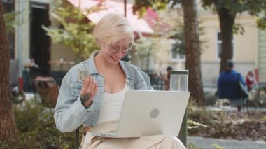 Mutlu sarışın kadın dizüstü bilgisayar kazananı zaferini kullanıyor. Elinde nakit para tutuyor. Açık havada sandalyede oturuyor. Genç kız turist yatırım bütçesi başarısı.