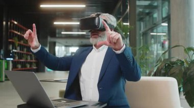 Olgun bir iş adamı yazılım geliştiricisi kulaklık uygulaması kullanarak sunum yapıyor. Sanal gerçeklik 3D 360 videosu izliyoruz. Ofis lobisinde çalışan, VR gözlüklü, beyaz saçlı, takım elbiseli adam..