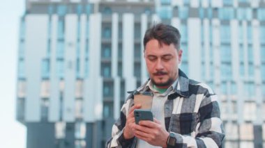 Mutlu adam akıllı telefon kullanıyor mesajlar yazıyor internet sosyal medya uygulamalarına bakıyor internette sohbet ediyor sabahın tadını çıkarıyor sıcak kahve içmek için dışarı çıkıyor. Şehir caddesinde yürüyen bir adam..