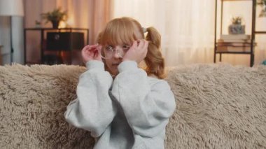 Genç bir kız evde düşünceli ve odaklanmış bir ifadeyle gözlük takıyor. Kafkasyalı çocuk kanepede oturuyor ve rahat oturma odasında günbatımında sakin ve pratik bir tavırla göz giysisini ayarlıyor.