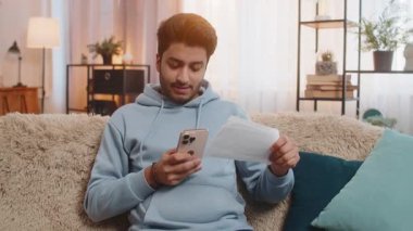 Uygulama yoluyla faturaları ödeyen genç Hintli adam elinde akıllı telefon ve makbuzlarla evdeki oturma odasındaki kanepede oturuyor. Hispanik adam doğru ödeme detaylarını özenle gözden geçiriyor..