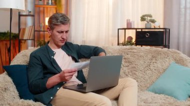 Evdeki genç adam kızgın görünüyor ve faturalarını kanepeye atarken bilgisayarını kapatıyor. Kafkasyalı erkek, bilgisayar sorunu veya mali belgelerin neden olduğu görülebilir hayal kırıklığı ve stresle tepki veriyor.