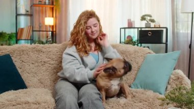 Genç mutlu Kafkasyalı kızıl saçlı kadın pug dog 'u koltukta nazikçe okşuyor, sıcakkanlılıkla ve yumuşak duygularla gülümsüyor. Evcil hayvan yakınımızda kalır, temastan zevk alır ve evde samimi bir dostluk içinde birlikte dinlenir..