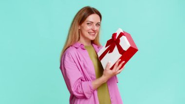 Orta yaşlı bir kadın ilginç bir şekilde bir hediye kutusunu açıyor ve içinde ne olduğuna mutlu bir şekilde tepki veriyor, heyecan, neşe ve şaşkınlık gösteriyor. Mavi arka planda izole edilmiş bir kız neşeli duygular ve hareketlerle kutluyor..