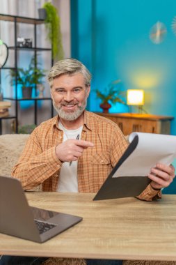 Beyaz, olgun bir adam evde oturmuş önemli belgeleri inceliyor ve dizüstü bilgisayar kullanıyor. Orta yaşlı bir adam başarılı bir işten aldığı memnuniyetle gülümser, verimlilik ve başarıyı gösterir.