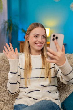 Beyaz bir kadın evdeki kanepede oturuyor, gülümsüyor, gülüyor, neşeli telefon görüşmesinde el sallıyor. Mutlu kız heyecan verici haberleri paylaşıyor, akıllı telefonu kullanıyor, evin içinde rahatça eğleniyor.