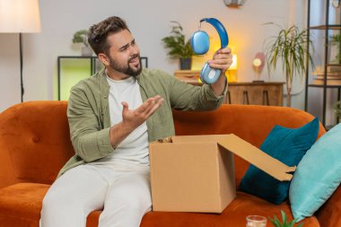 Mutsuz genç beyaz adam kutuları açıyor, kablosuz kulaklık siparişinden memnun değil, mobil uygulamalara olumsuz geri bildirim yazıyor, internet alışverişi kavramı.